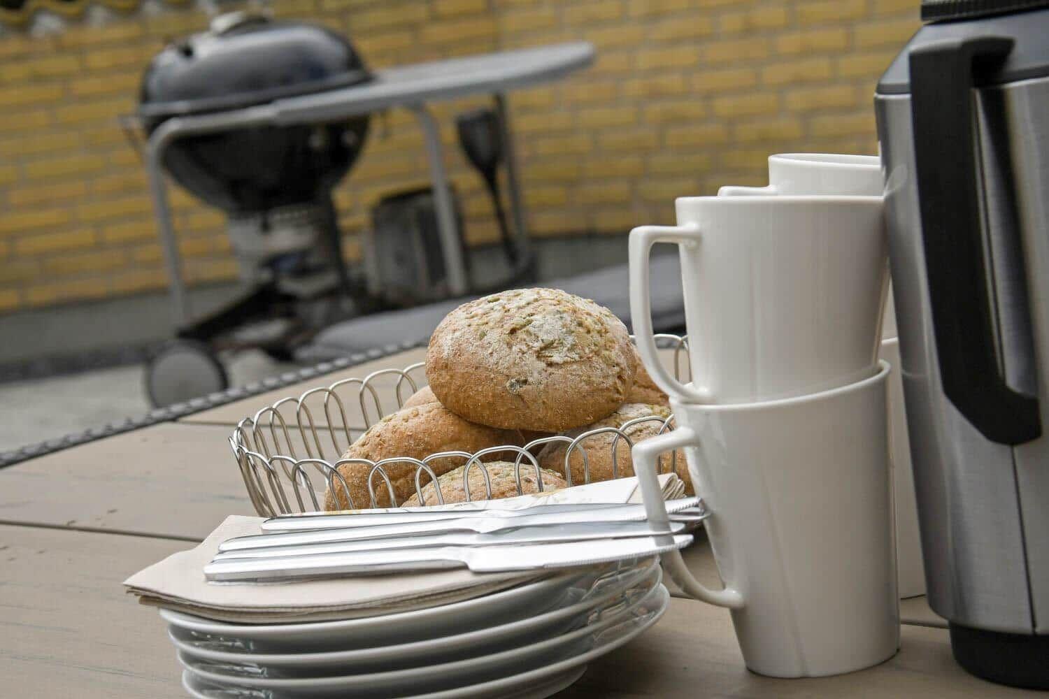 Tisch mit Brot, Tassen und Geschirr vor Grill und gelbem Mauerwerk.