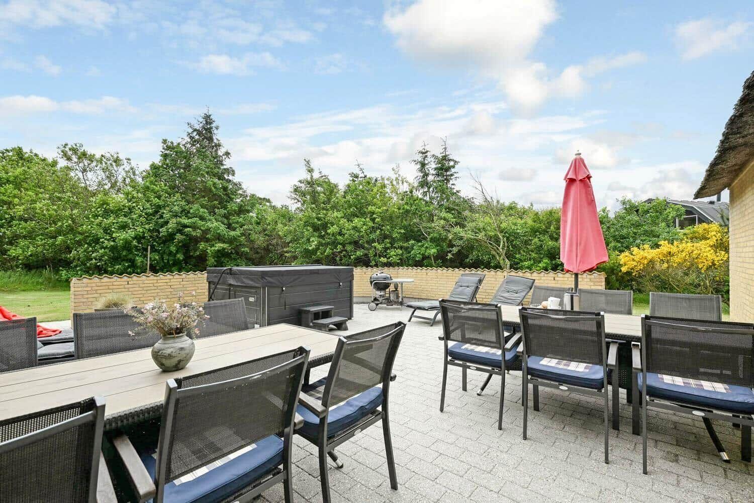 Terrasse mit Tisch, Stühlen, Sonnenschirm und Grill. Hintergrund: Grünflächen und Hecke.