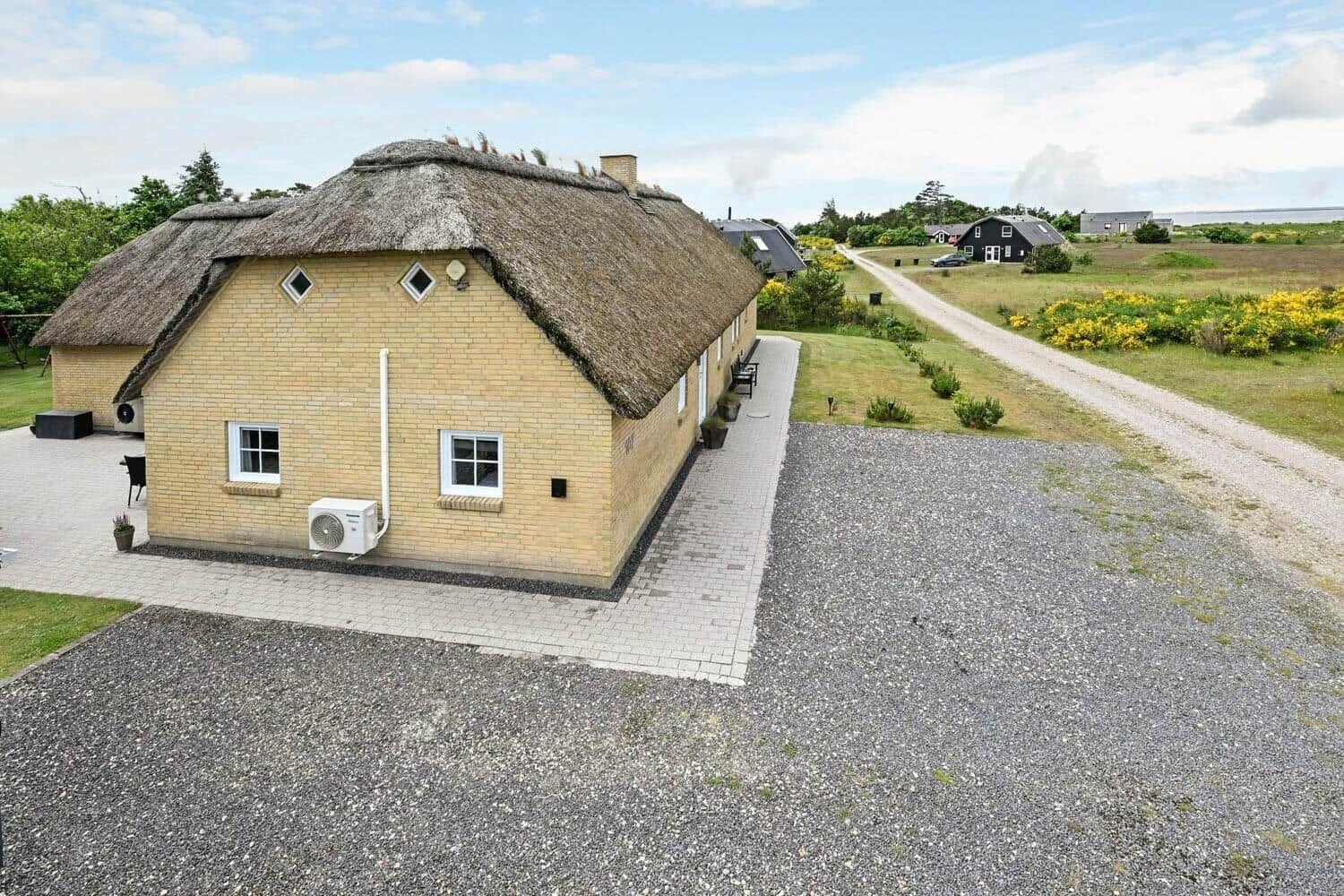 Haus mit strohgedecktem Dach, gepflasterter Weg und Kiesfläche. Grünflächen und Straße im Hintergrund.