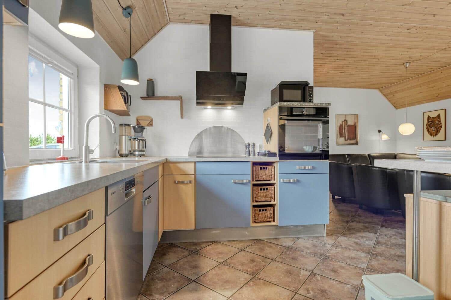 Küche mit Holzdecke, blauen Schränken und Steinplattenboden. Esstisch mit schwarzen Stühlen.