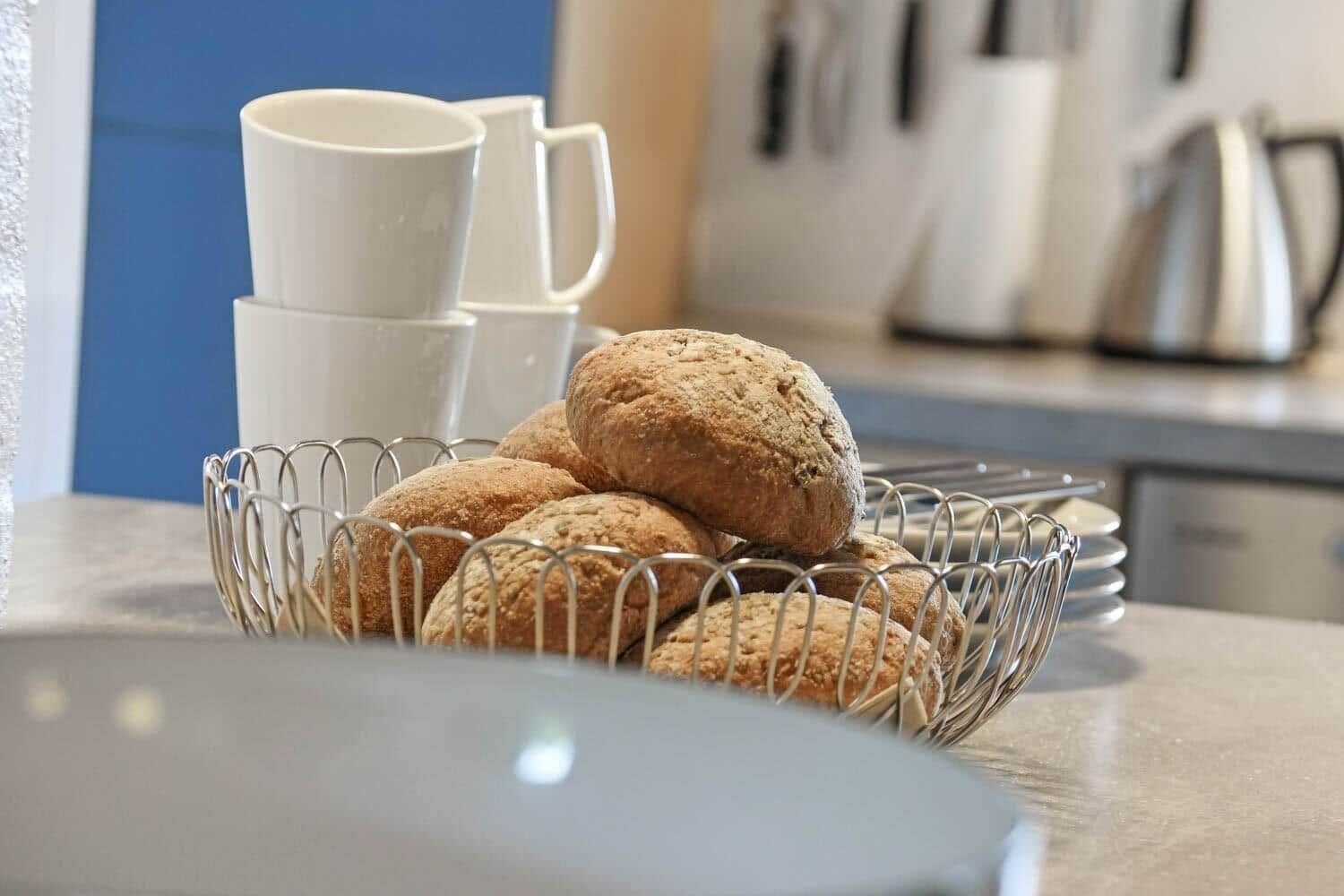 Küchenarbeitsplatte mit Korb voller Brötchen und weißen Tassen.