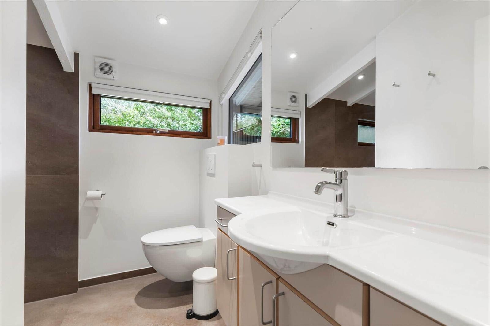 Bathroom with sink, mirror, toilet, and window with view of greenery.