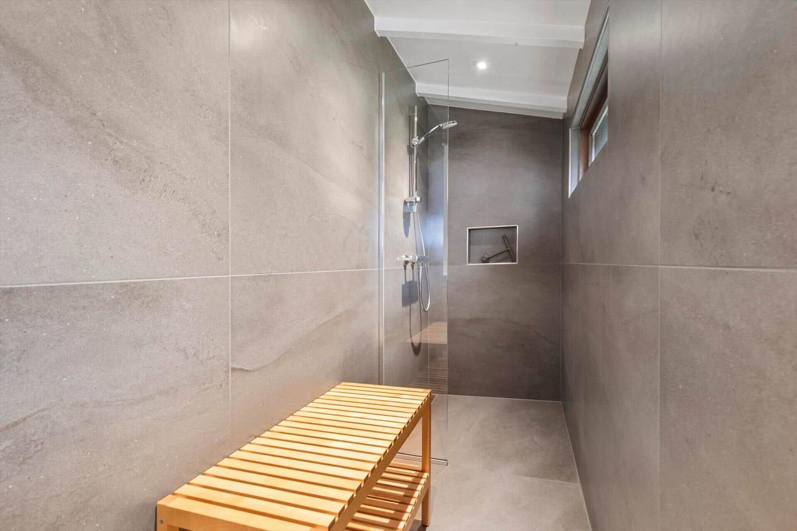 Shower area with glass enclosure, wooden bench, and wall niche. Floor and walls are tiled with stone.