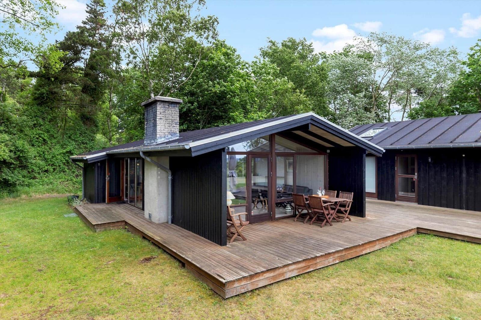 Modern villa with wooden terrace and garden. Large glass front, seating areas, and barbecue space.
