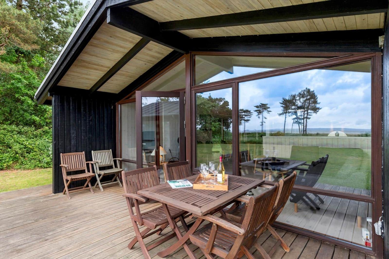 Deck with wooden table and chairs, large glass walls, view of lawn and trees.