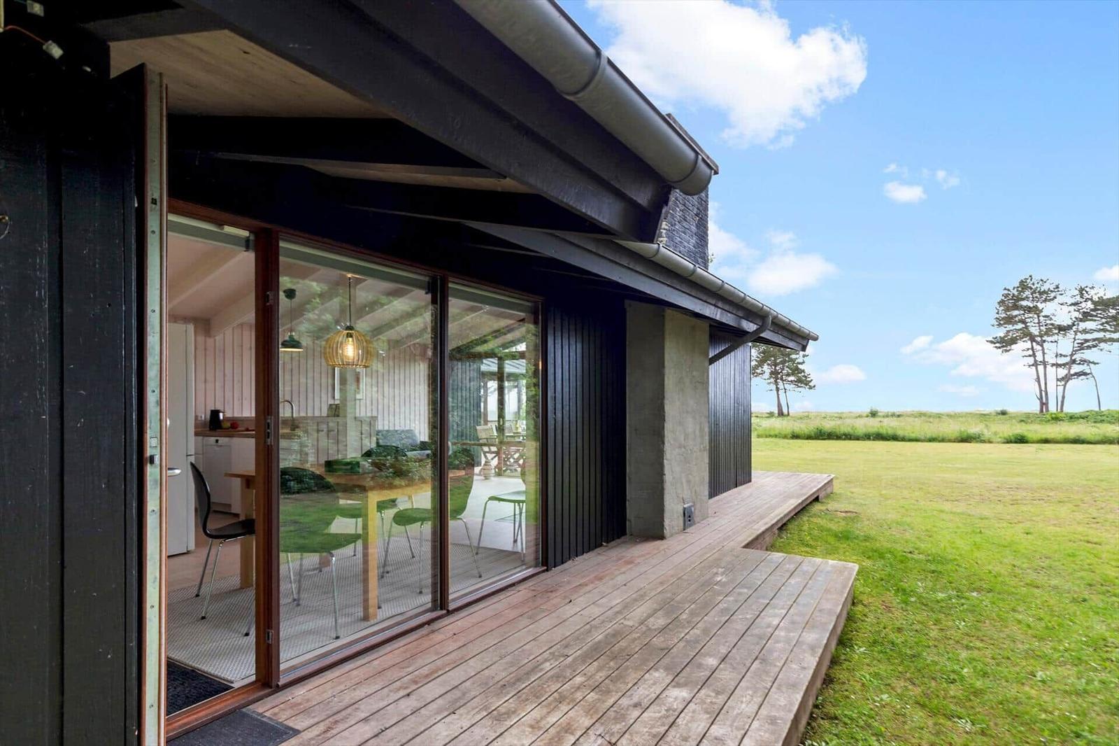 Modern villa with wooden terrace and large glass area. View of green field and trees.
