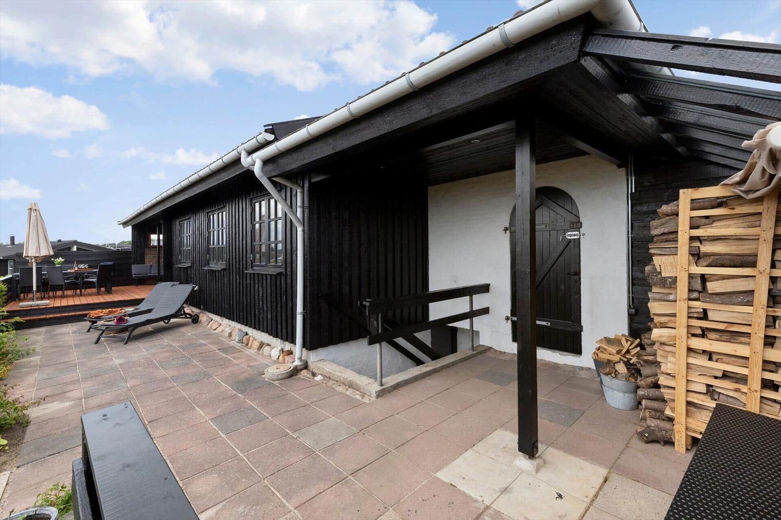 Schwarzes Holzhaus mit Terrasse, Liegestühlen und Holzstapel. Helle Fenster und Dachüberstand.