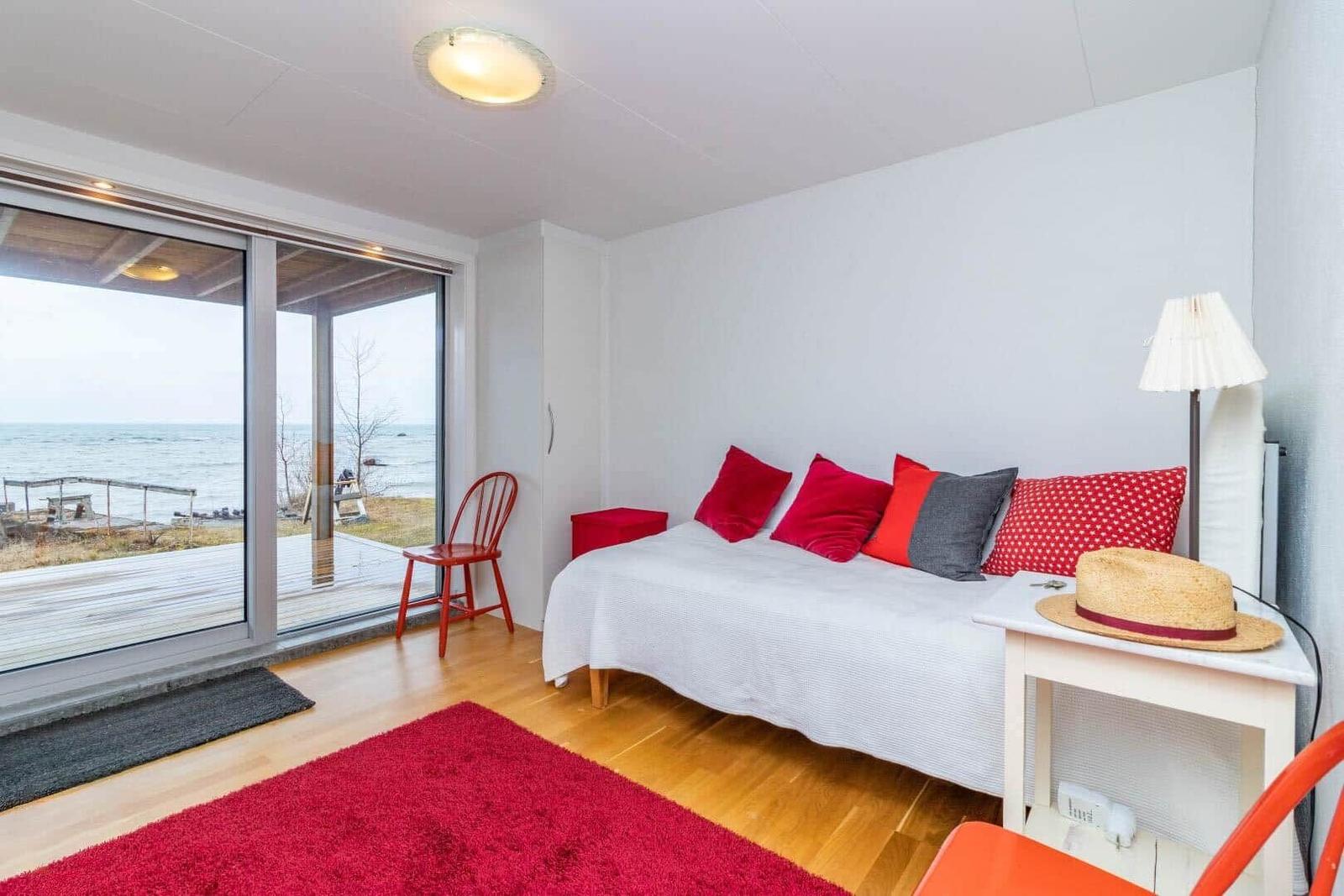 Schlafzimmer mit Blick aufs Meer, rote Polster und Teppich, weiße Wand und Holzboden.
