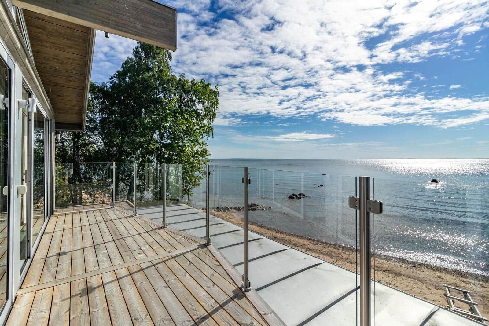 Terrasse mit Holz- und Betonfläche, Glasgeländer und Blick auf See und Strand.