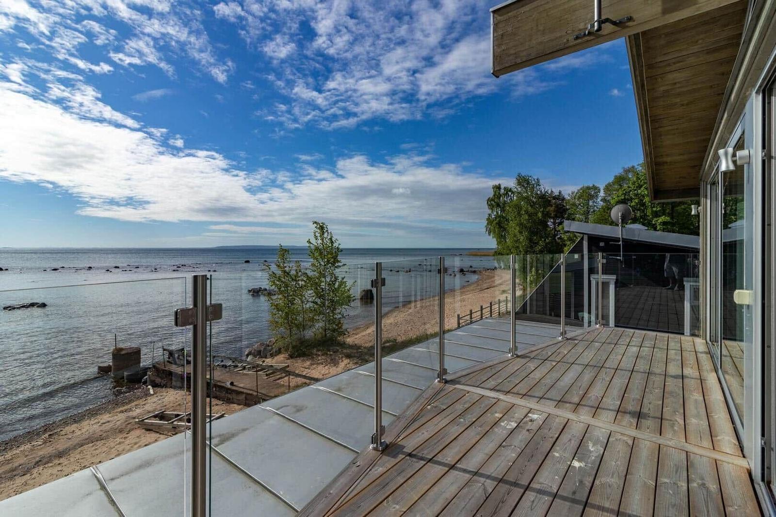 Terrasse mit Holz- und Glasgeländer überblickt einen Strand und See unter blauem Himmel.