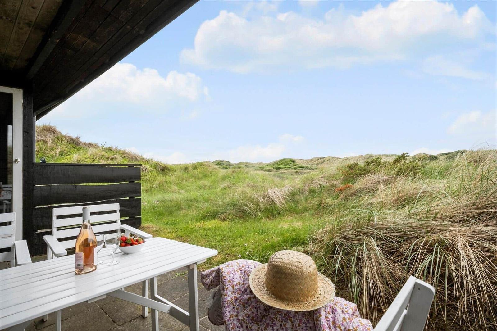 Eine Person sitzt auf einer Terrasse mit Tisch und Stühlen. Im Hintergrund sind Dünen und Gras sichtbar.
