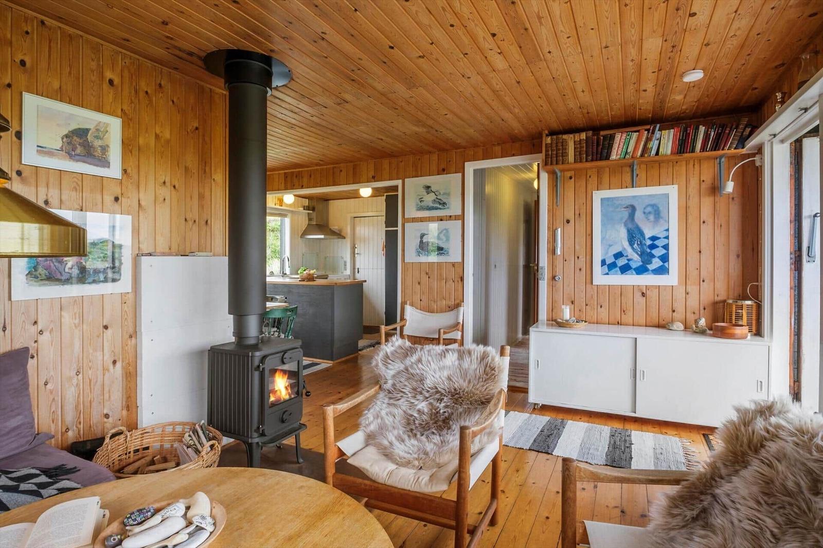 Wohnraum mit Kaminofen, Holzwänden und Sitzgelegenheiten. Küche im Hintergrund.