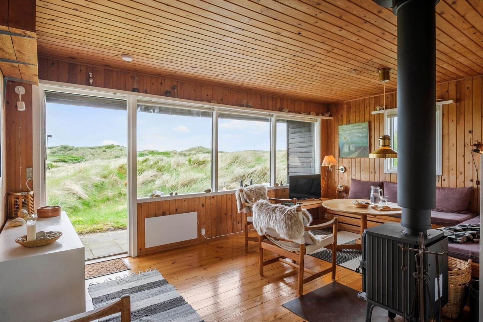 Wohnzimmer mit Holzwand, Kamin und großem Fensterblick auf Dünen.