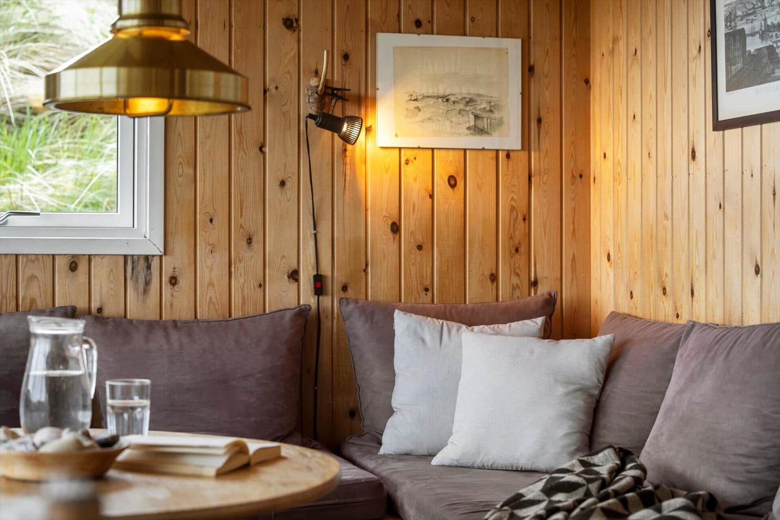 Ein Raum mit Holzwänden, Sofa, Tisch und Lampe. Ein Fenster zeigt Gras draußen.