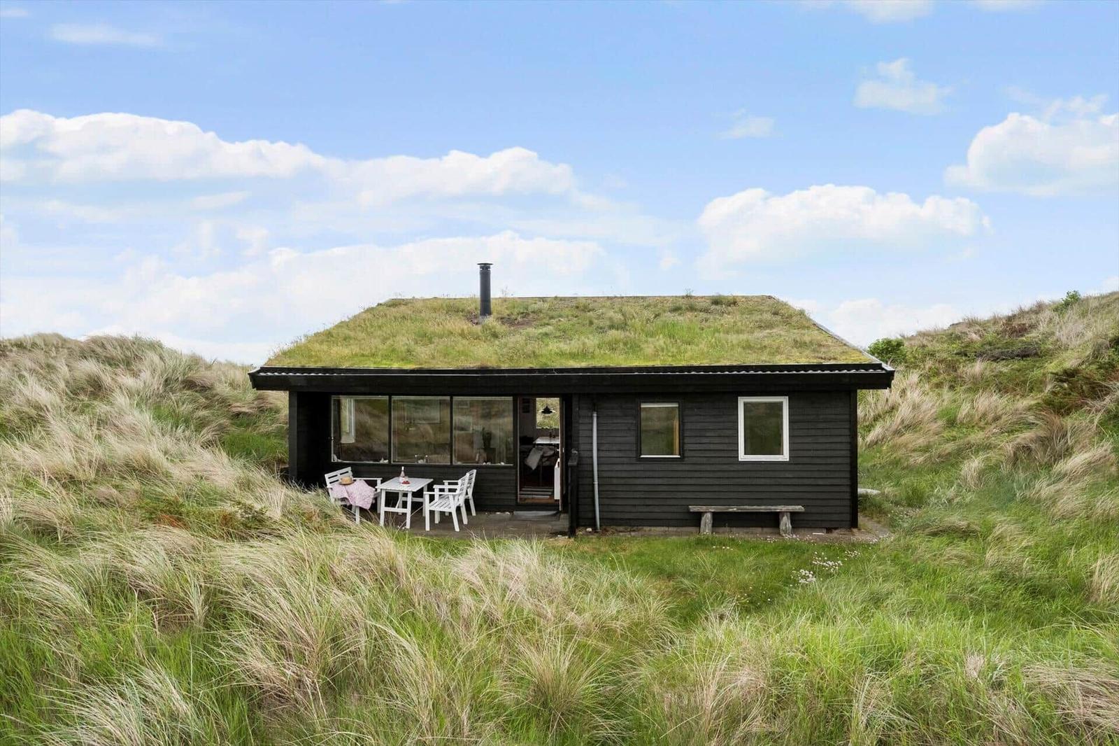 Schwarze Hütte mit Grasdach, Terrasse und Sitzgelegenheit zwischen Grasflächen.