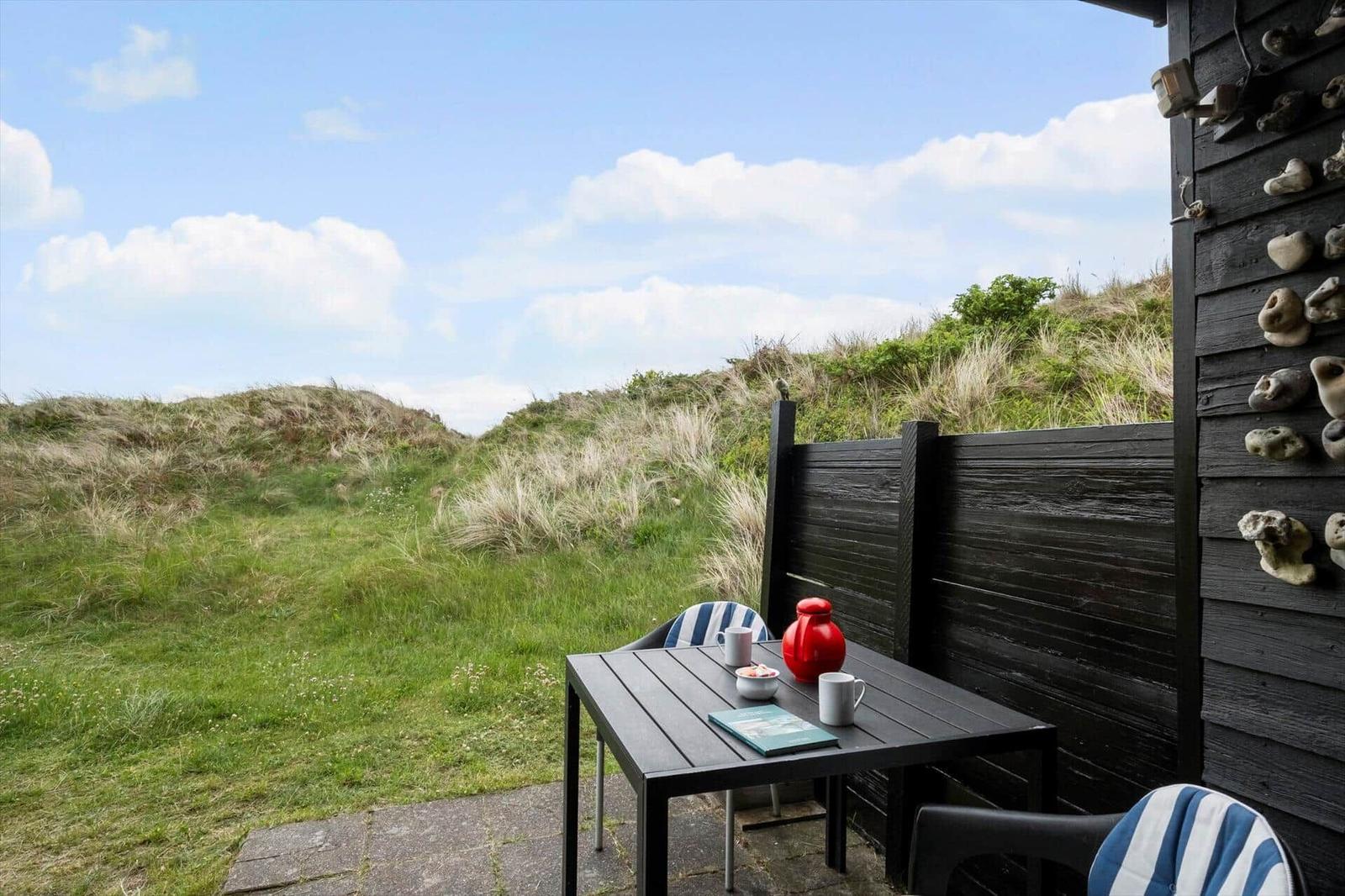 Terrasse mit Tisch und Stühlen, Blick auf grüne Dünen und Himmel.