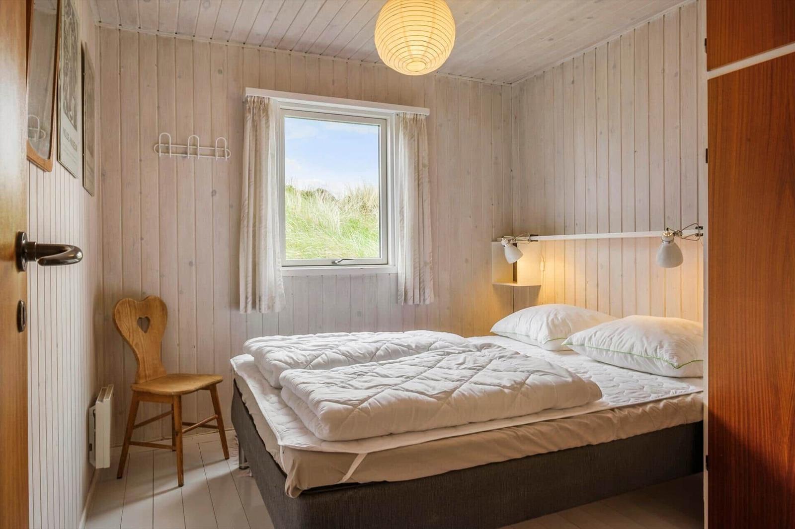 Schlafzimmer mit Bett, Fenster und Holzwand. Hängelampe und Stuhl sind sichtbar.