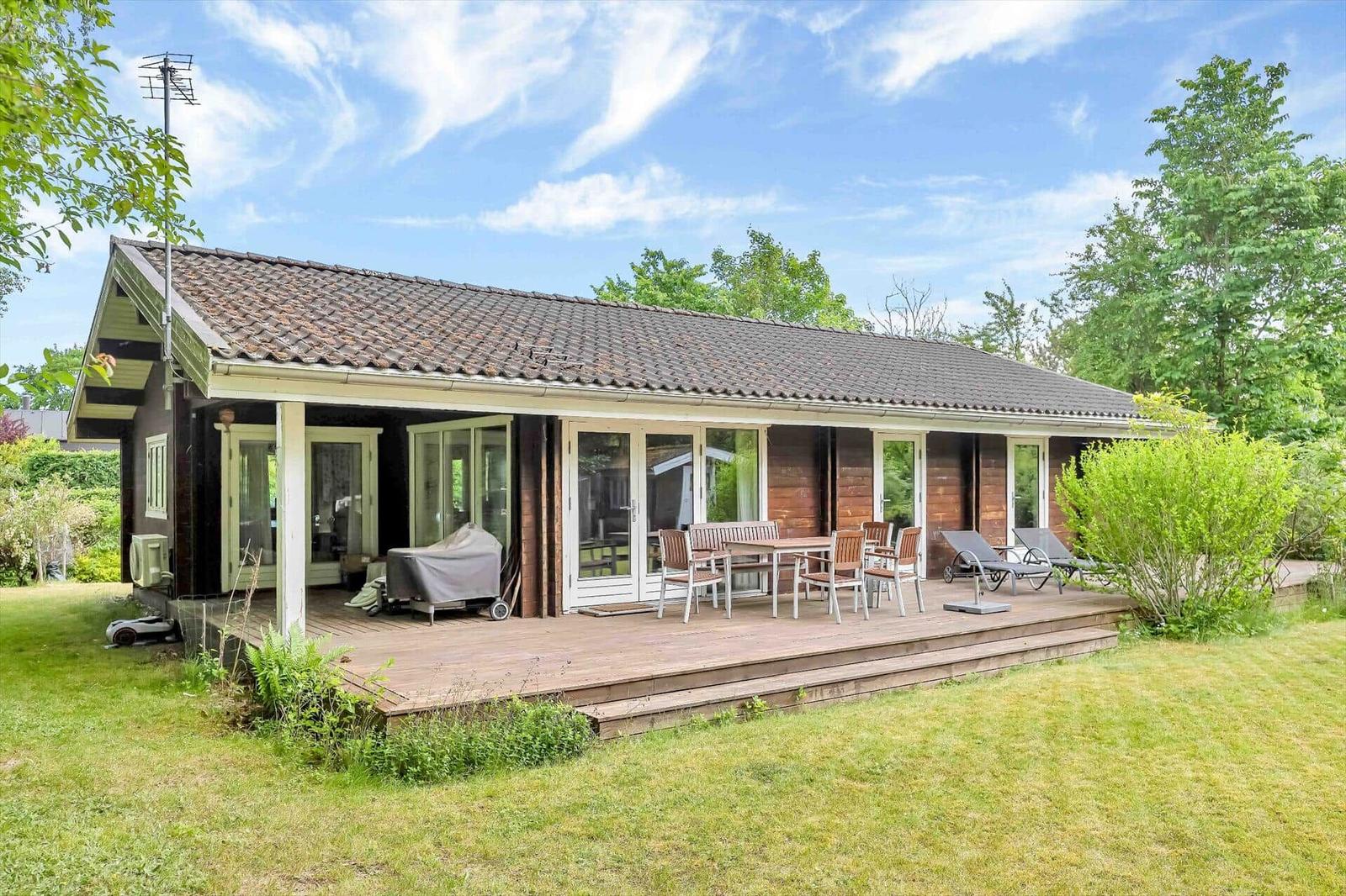 House with terrace, table, chairs, and loungers. Surrounded by green. Blue sky with clouds.