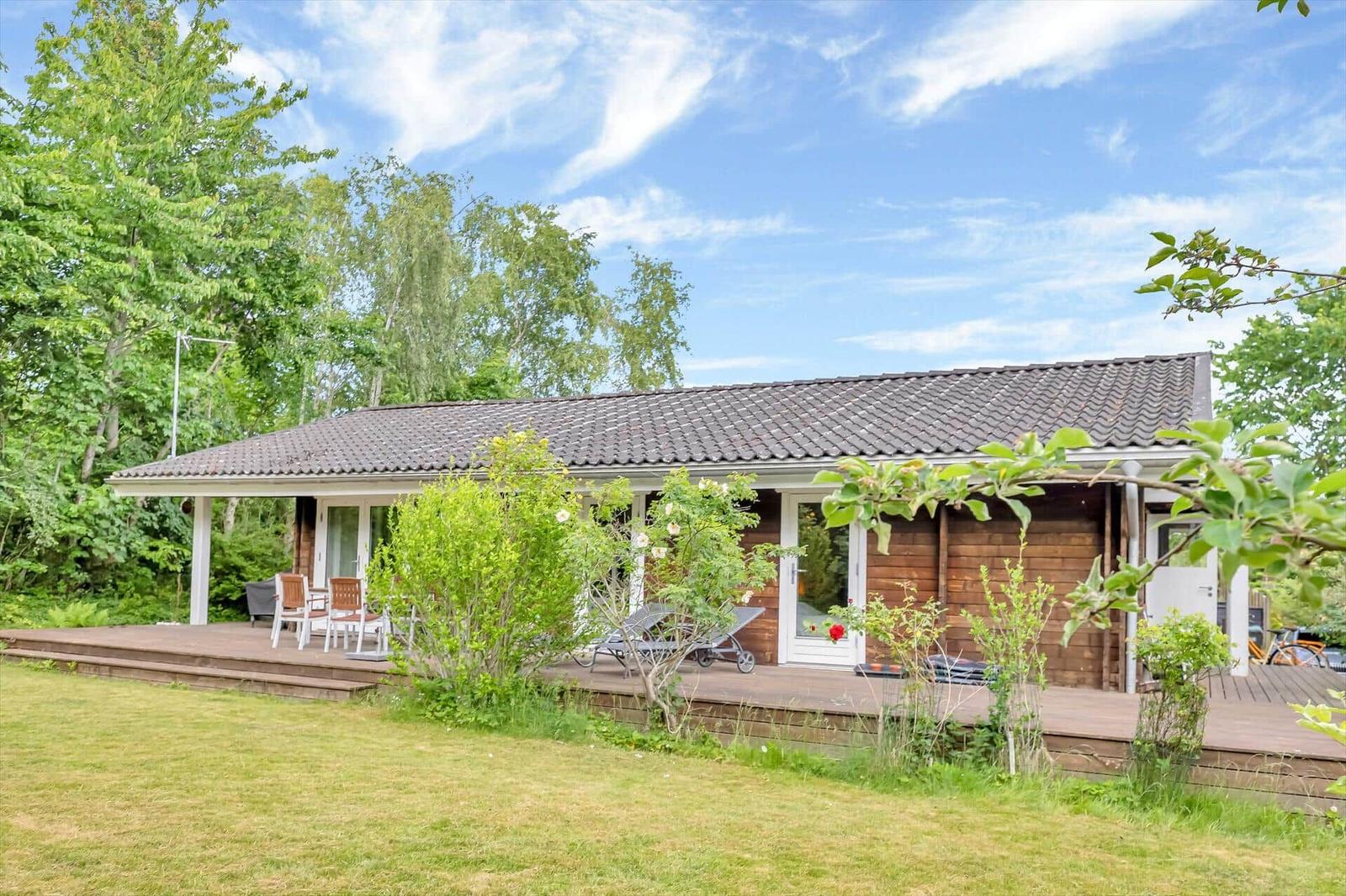 Wooden house with terrace, garden, and view of trees under blue sky.