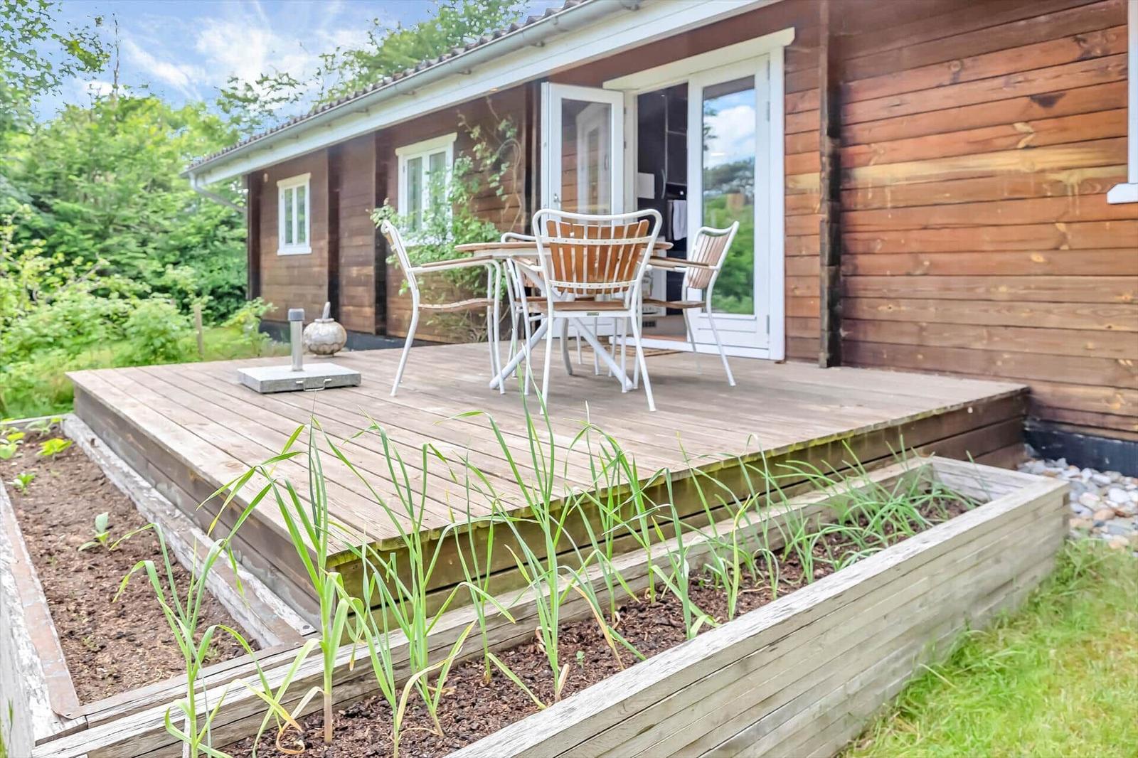 Deck with table and chairs in front of a wooden house with raised garden beds.