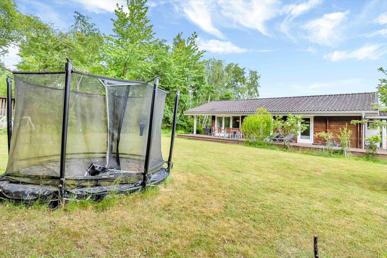 House with terrace, garden, and trampoline. Surrounded by trees under blue sky.