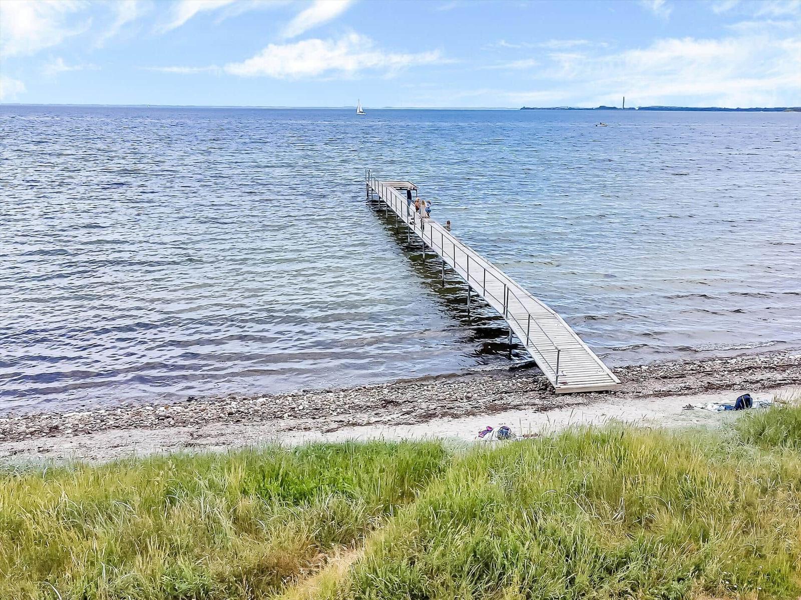 Long pier extends into the sea. People stand there. Beach and grass in the foreground.