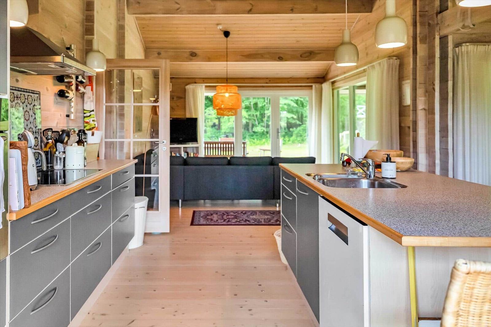 Kitchen with countertop, sink, and cabinets. In the background, living room with sofa and windows.