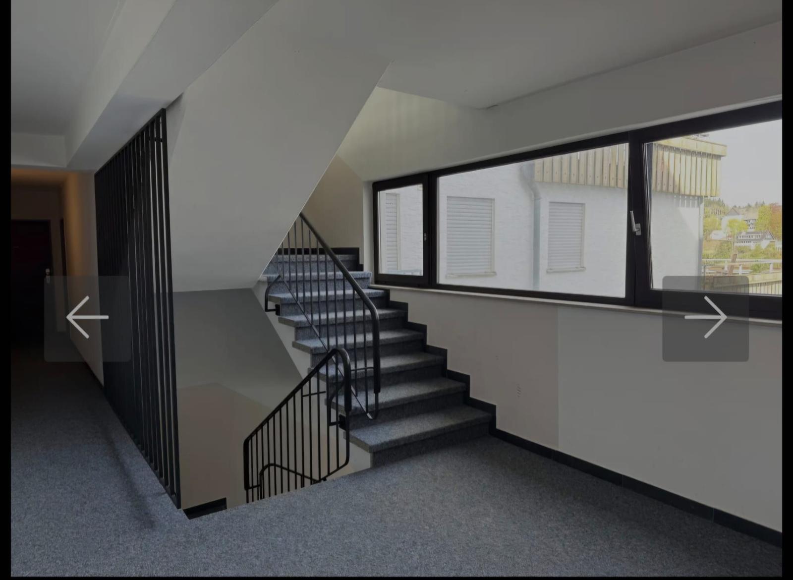 Treppe mit Geländer führt zu oberen Etagen. Große Fenster bieten Blick auf Außenbereich.