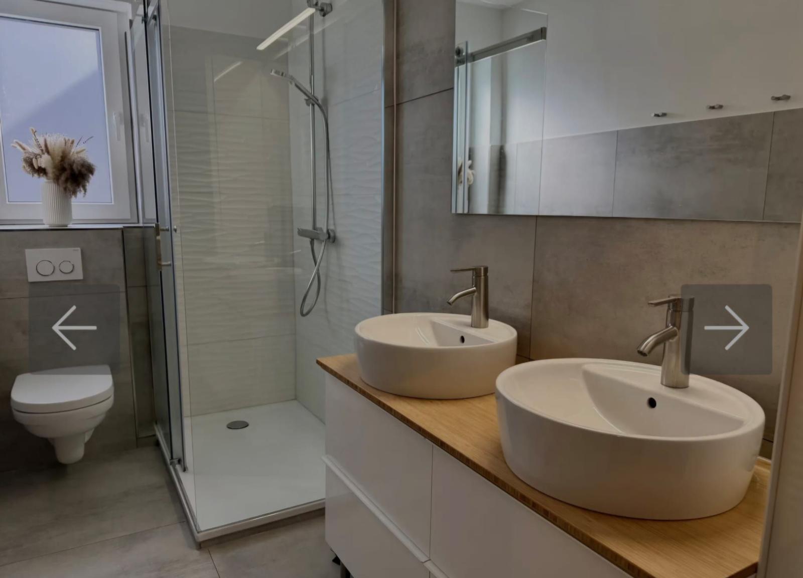 Badezimmer mit Dusche, Toilette und zwei Waschbecken auf Holzplatte.