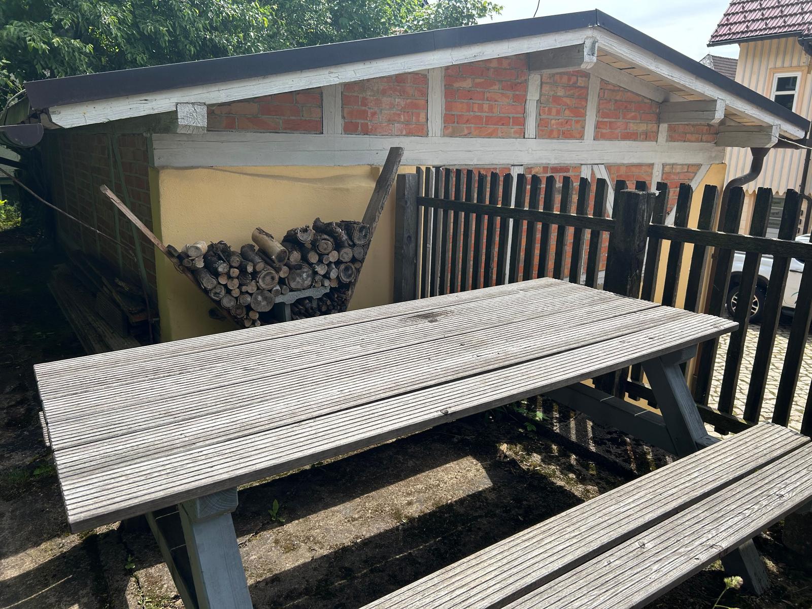 Holztisch mit Bänken im Garten vor einem Schuppen mit Feuerholz.