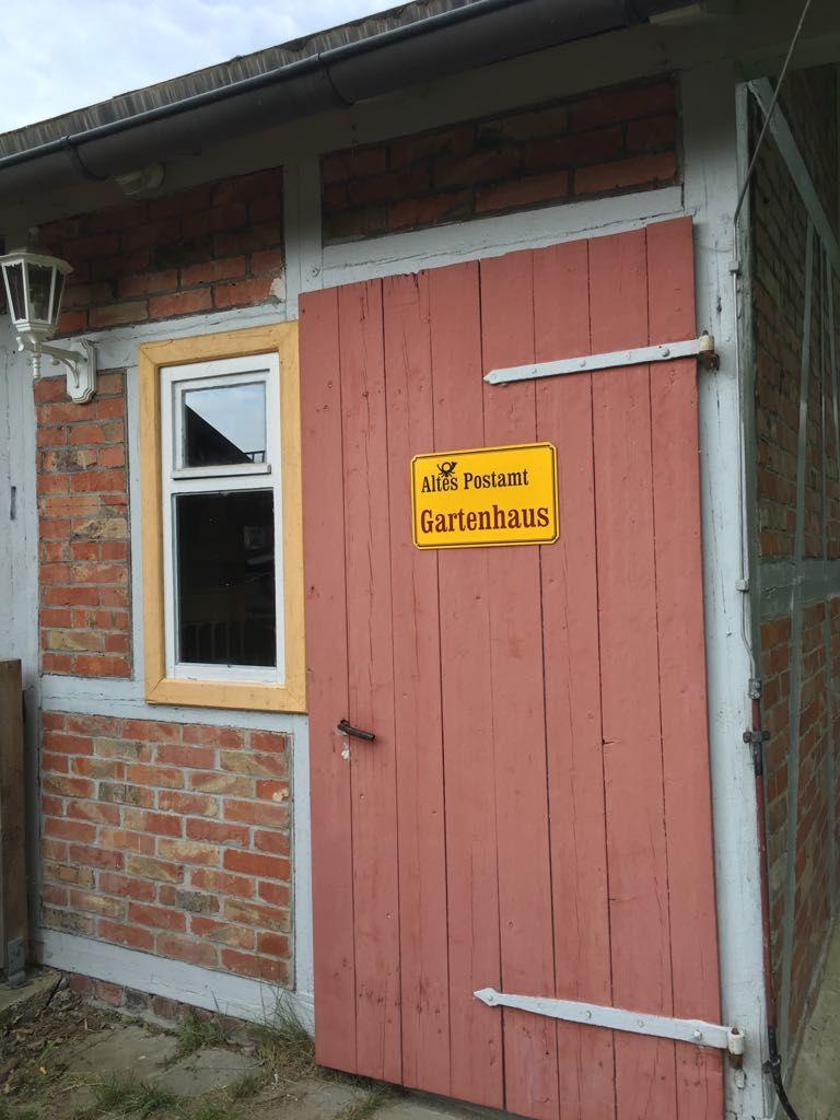 Rotes Tor mit Schild Altes Postamt Gartenhaus an Backsteinhaus mit Fenster.