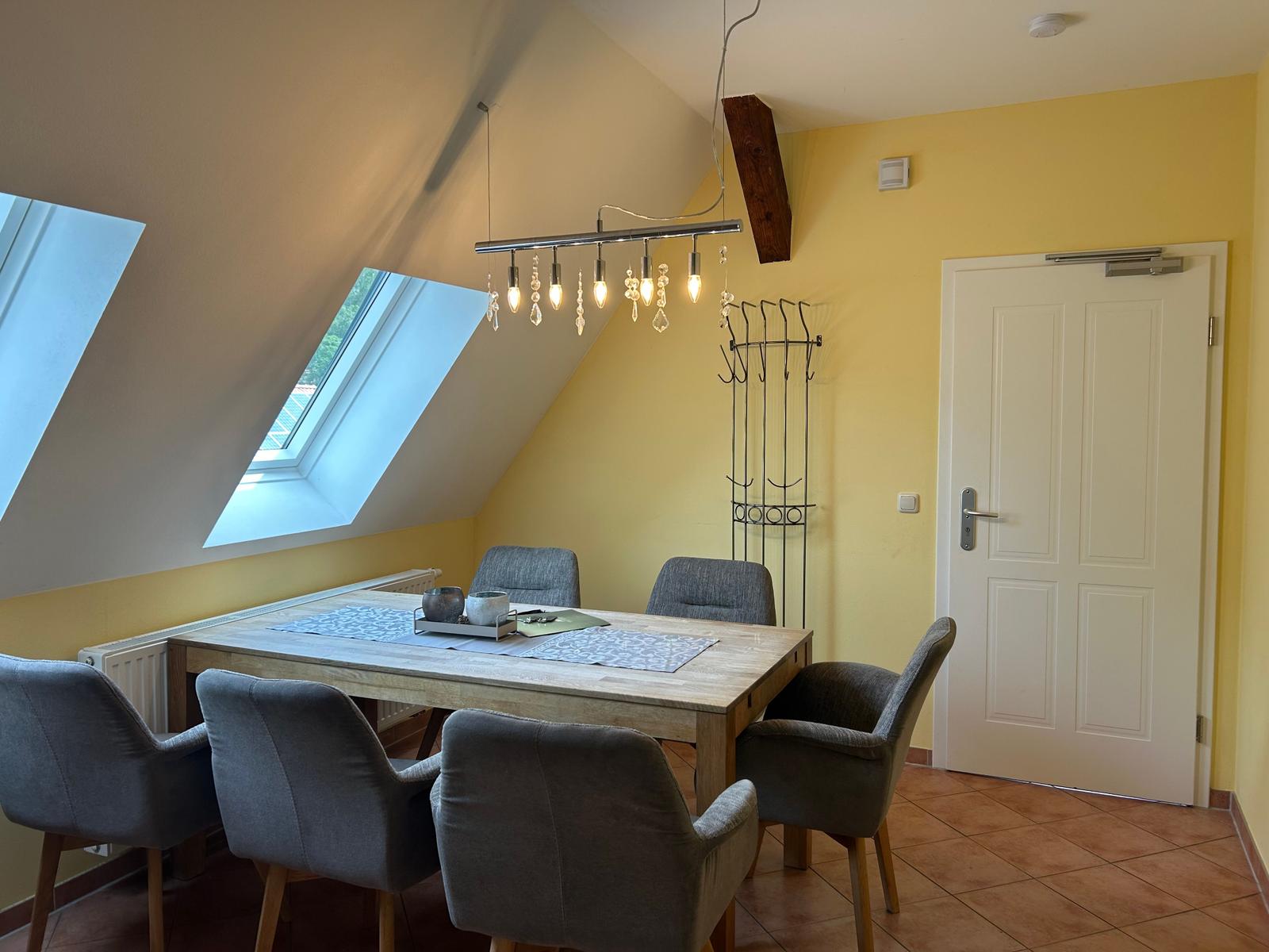 Esszimmer mit Holztisch, sechs Stühlen und Dachfenstern. Gelbe Wände und Bodenfliesen.