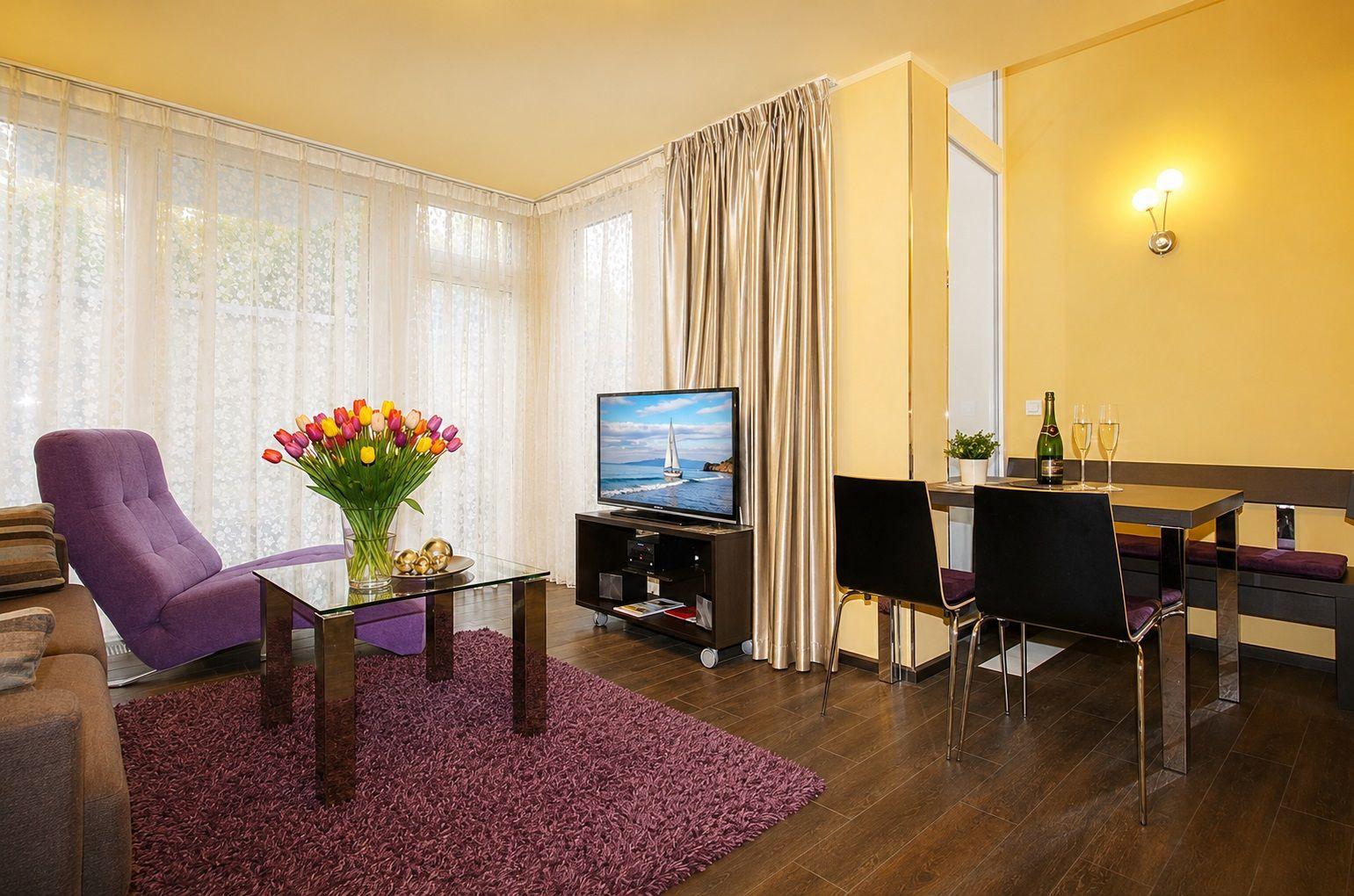 Wohnzimmer mit TV, Esstisch und lila Sitzmöbel vor großen Fenstern.