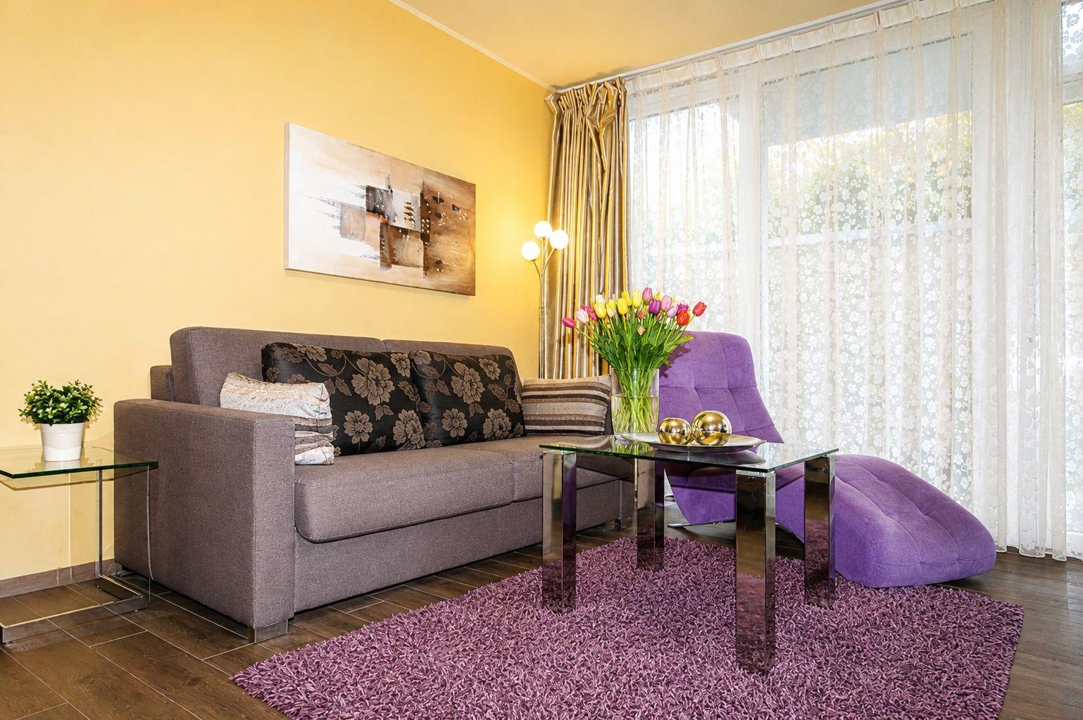 Wohnzimmer mit Sofa, Vase mit Tulpen und lila Sessel vor Fenster mit Vorhängen.