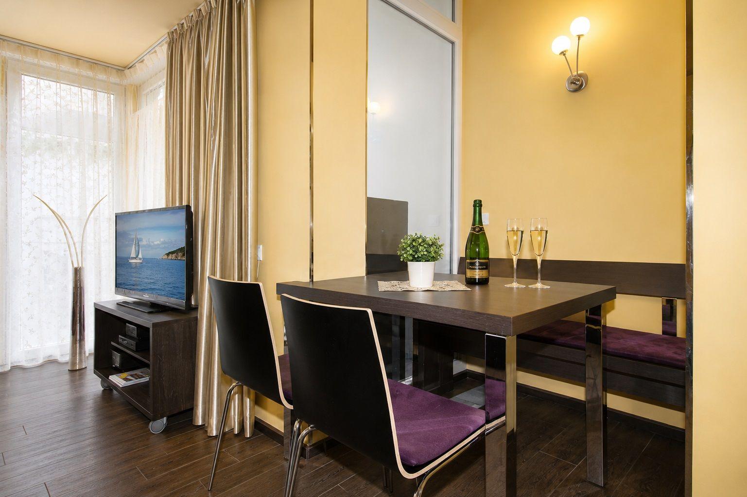 Essbereich mit Tisch, Stühlen und Flasche Champagner. Fernseher und Fenster mit Vorhängen.