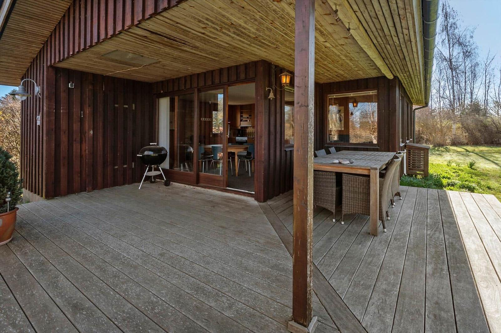 Terrasse mit Grill und Esstisch unter Holzüberdachung. Blick ins Haus durch Glastüren.