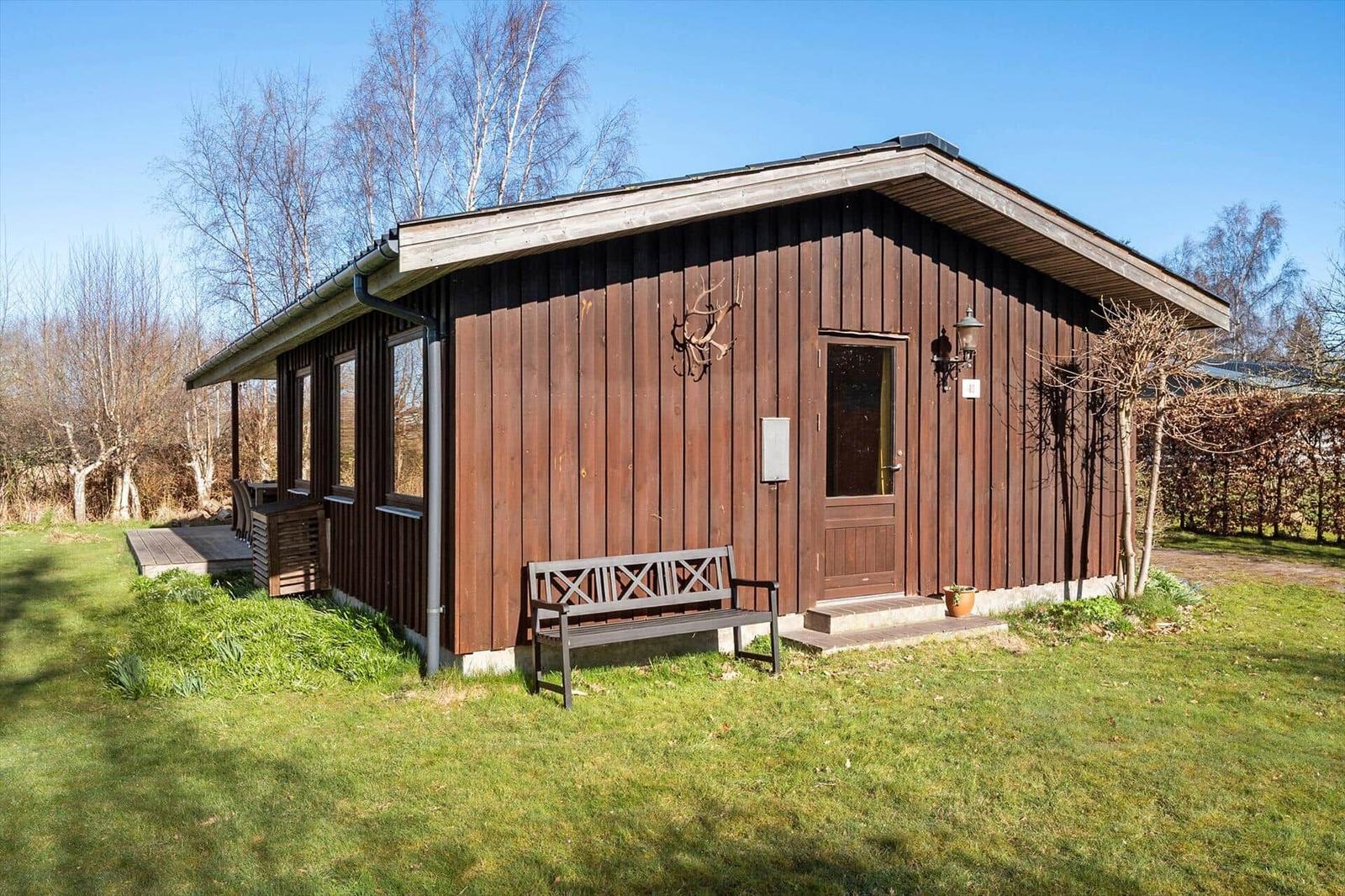 Holzhaus mit Eingangstür, Bank und Garten. Bäume im Hintergrund.