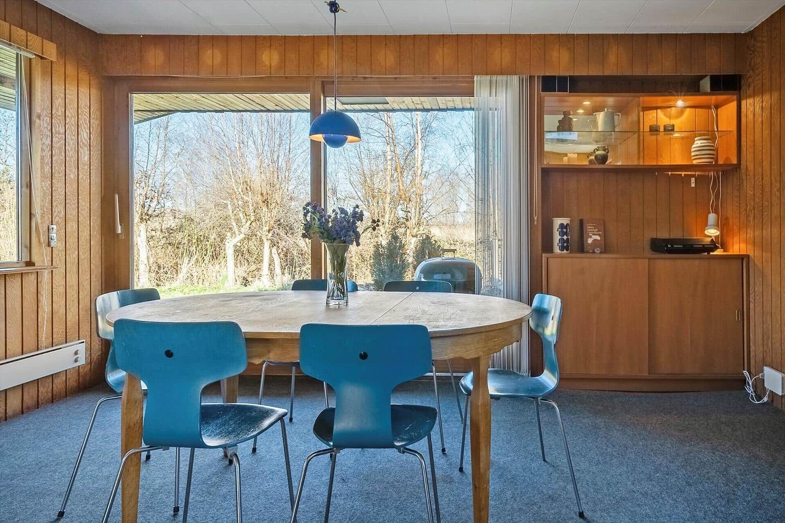 Esszimmer mit runden Tisch, blauen Stühlen und Holzwandverkleidung. Große Fenster mit Blick auf Außenbereich.