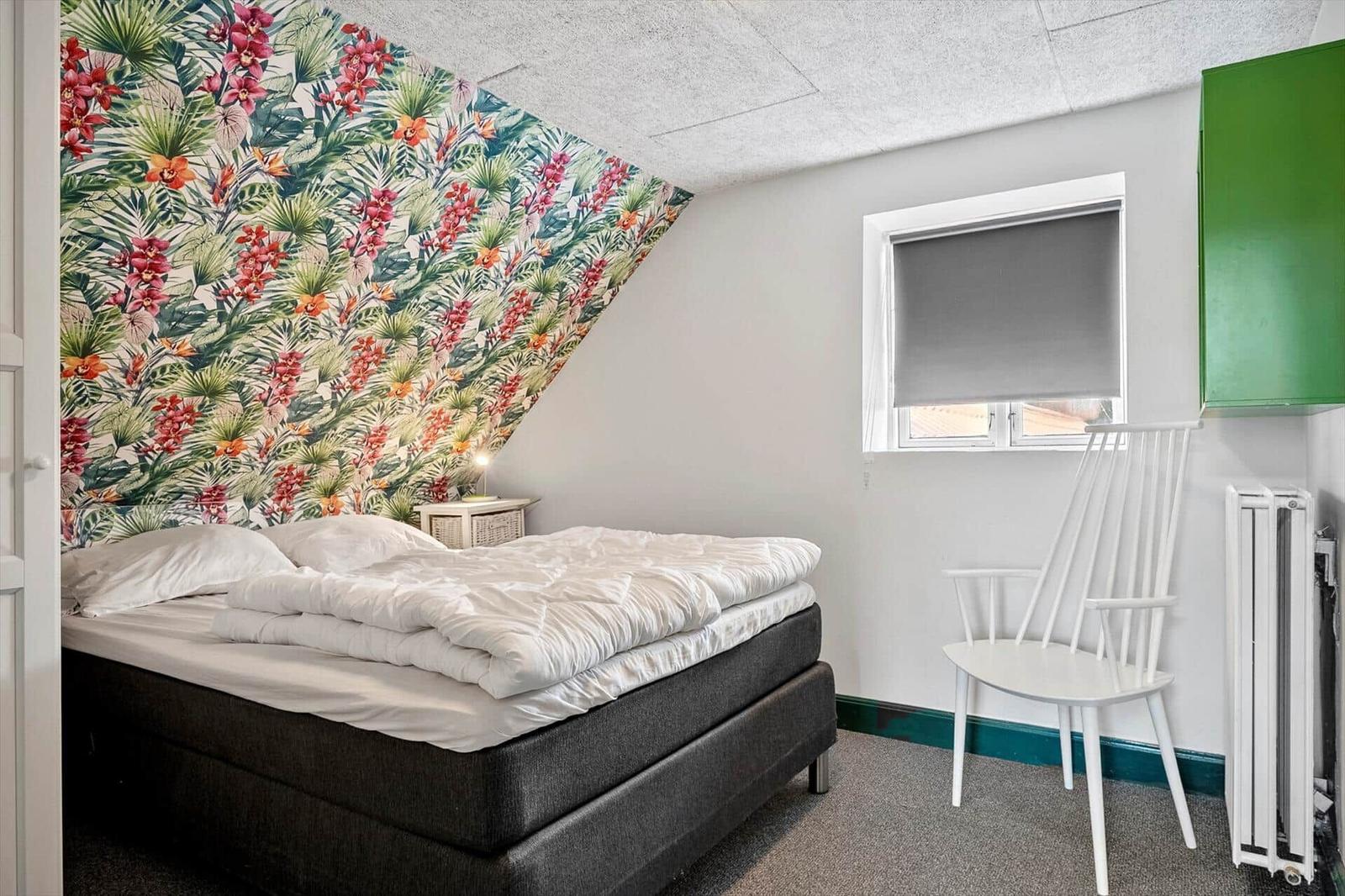 Schlafzimmer mit Bett, Stuhl und floraler Wandtapete. Fenster mit Rollladen.
