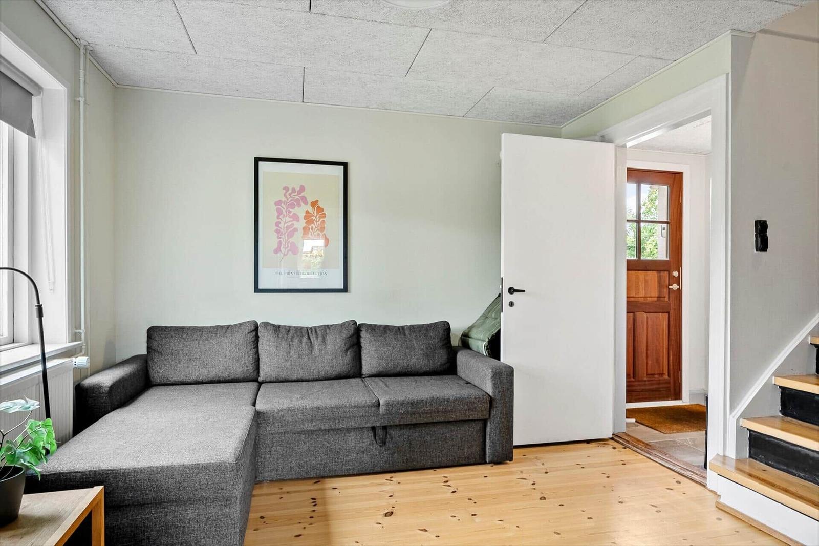 Wohnzimmer mit grauem Sofa, Holzboden und Wandbild. Tür führt ins Haus.