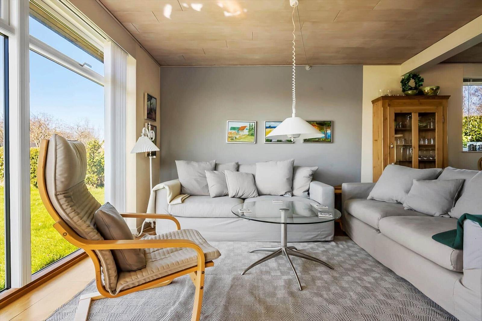 Wohnzimmer mit zwei Sofas, Glas-Tisch und Holzdecke. Große Fenster mit Blick auf Garten.