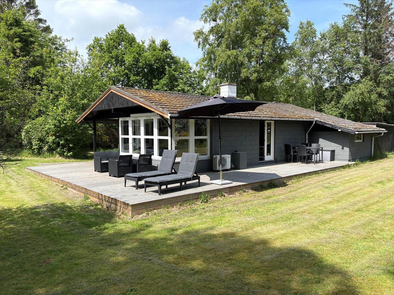 Haus mit Holzterrasse, Liegestühlen und Sonnenschirm. Umgeben von Gras und Bäumen.