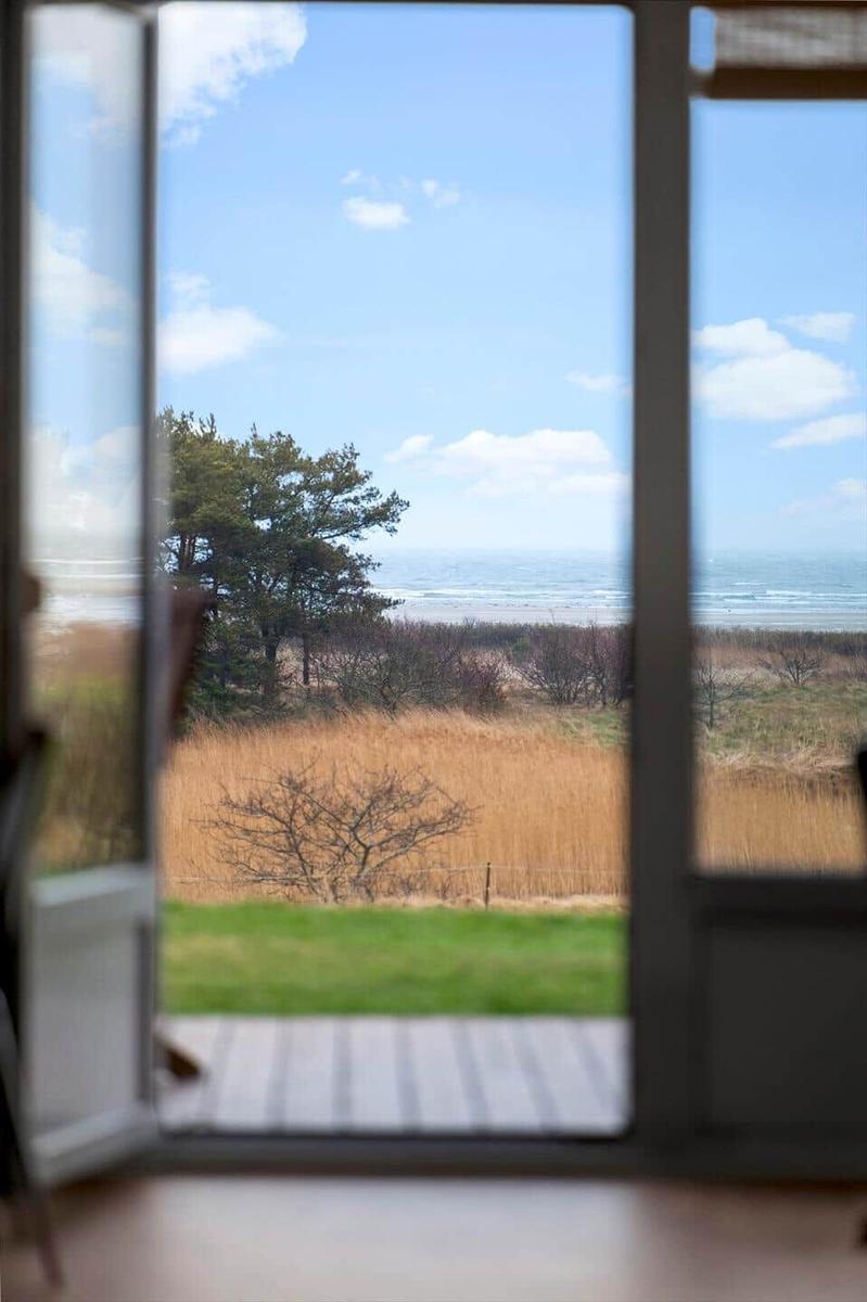 Durch die Glastür blickt man auf einen Strand mit Grasland und Bäumen unter blauem Himmel.