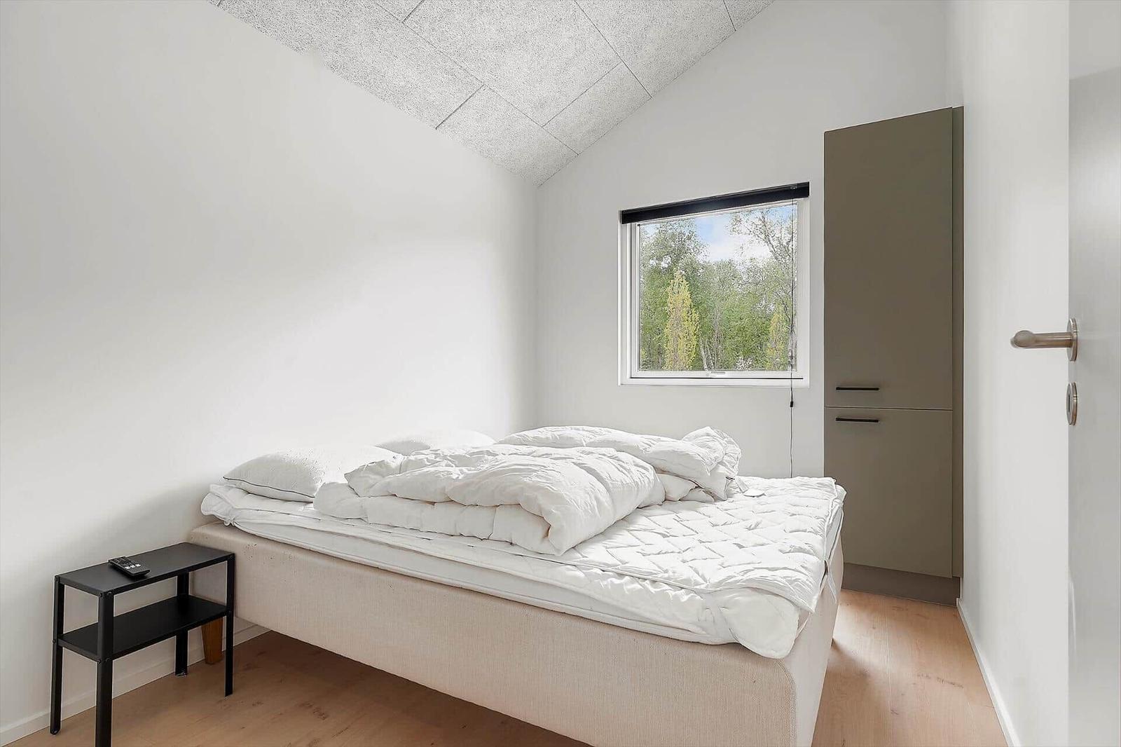 Schlafzimmer mit Bett, Nachttisch und Kleiderschrank. Fenster zeigt Baumwipfel.