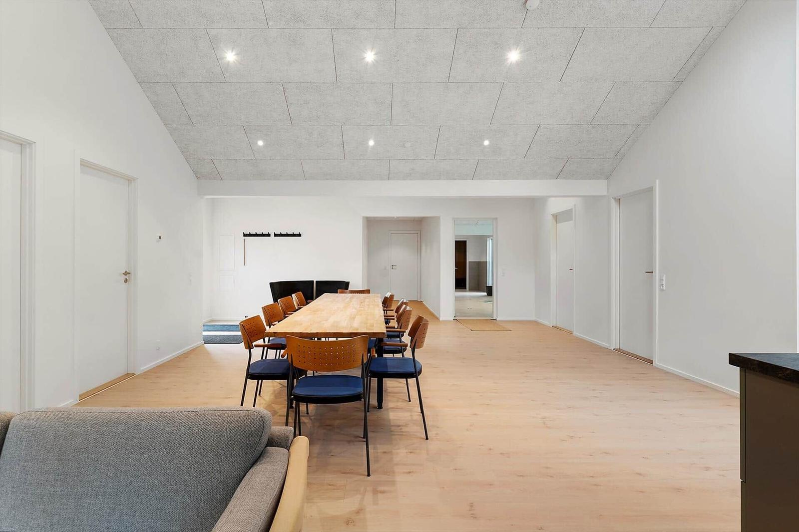 Großer Raum mit Holzboden, langem Tisch und Stühlen. Weiße Wände und Decke mit eingebauten Lichtern.