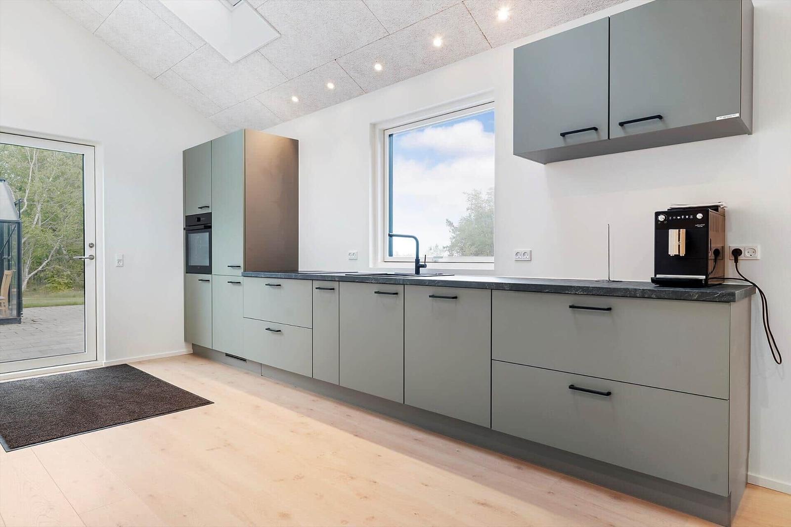 Moderne Küche mit grauen Schränken, dunkler Arbeitsplatte und Fenster mit Blick auf Außengelände.