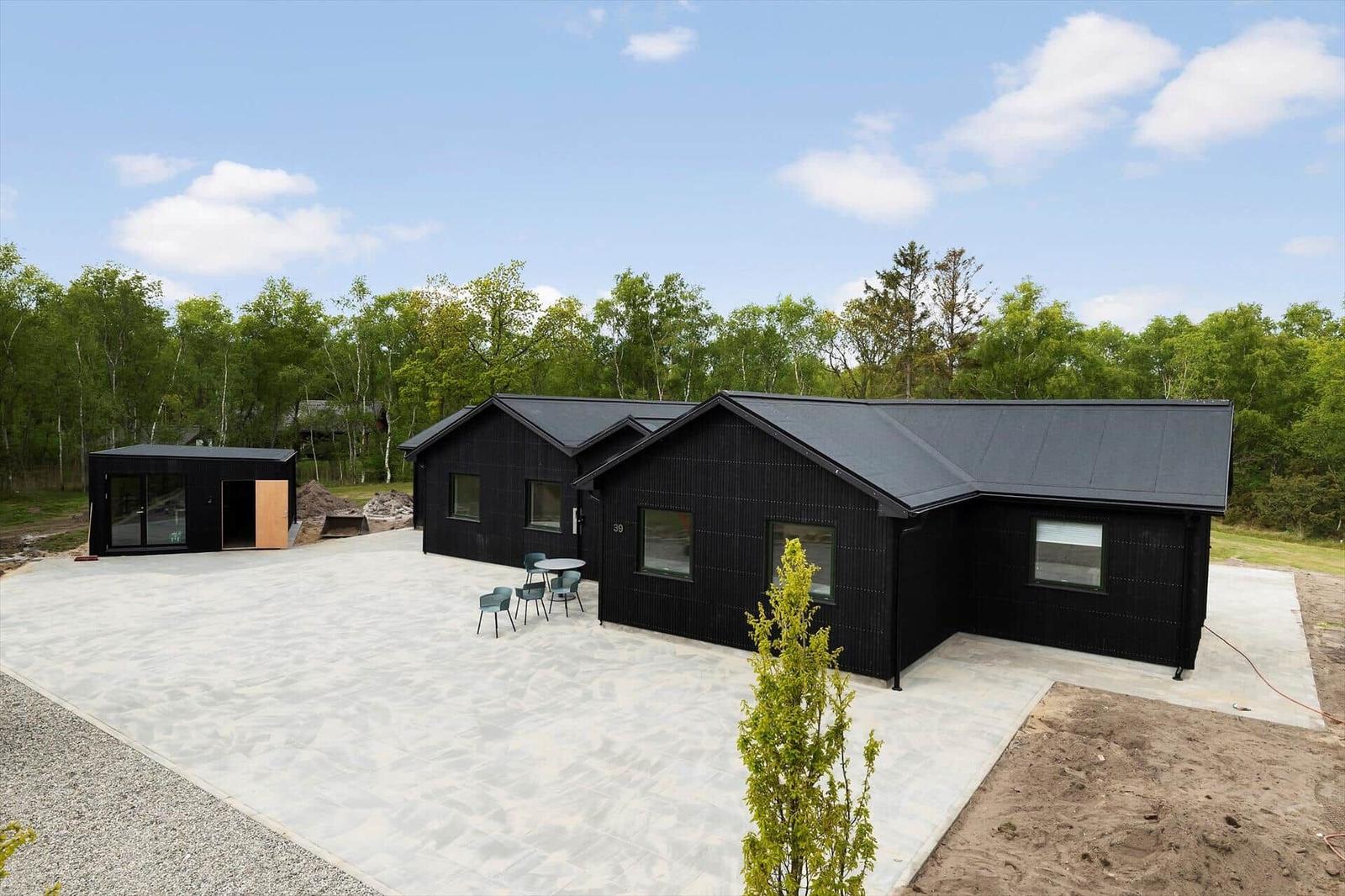 Schwarze Holzhütte mit Terrasse und Garten. Umgeben von Bäumen unter blauem Himmel.