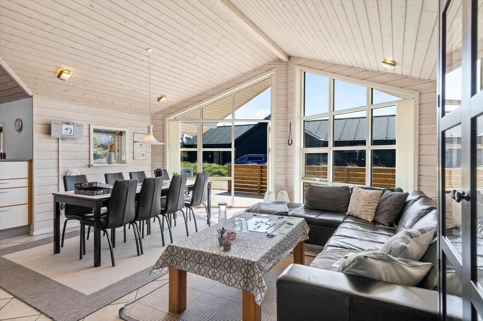 Wohn- und Essbereich mit Sofa, Tisch und Stühlen. Große Fensterfront mit Blick nach draußen.