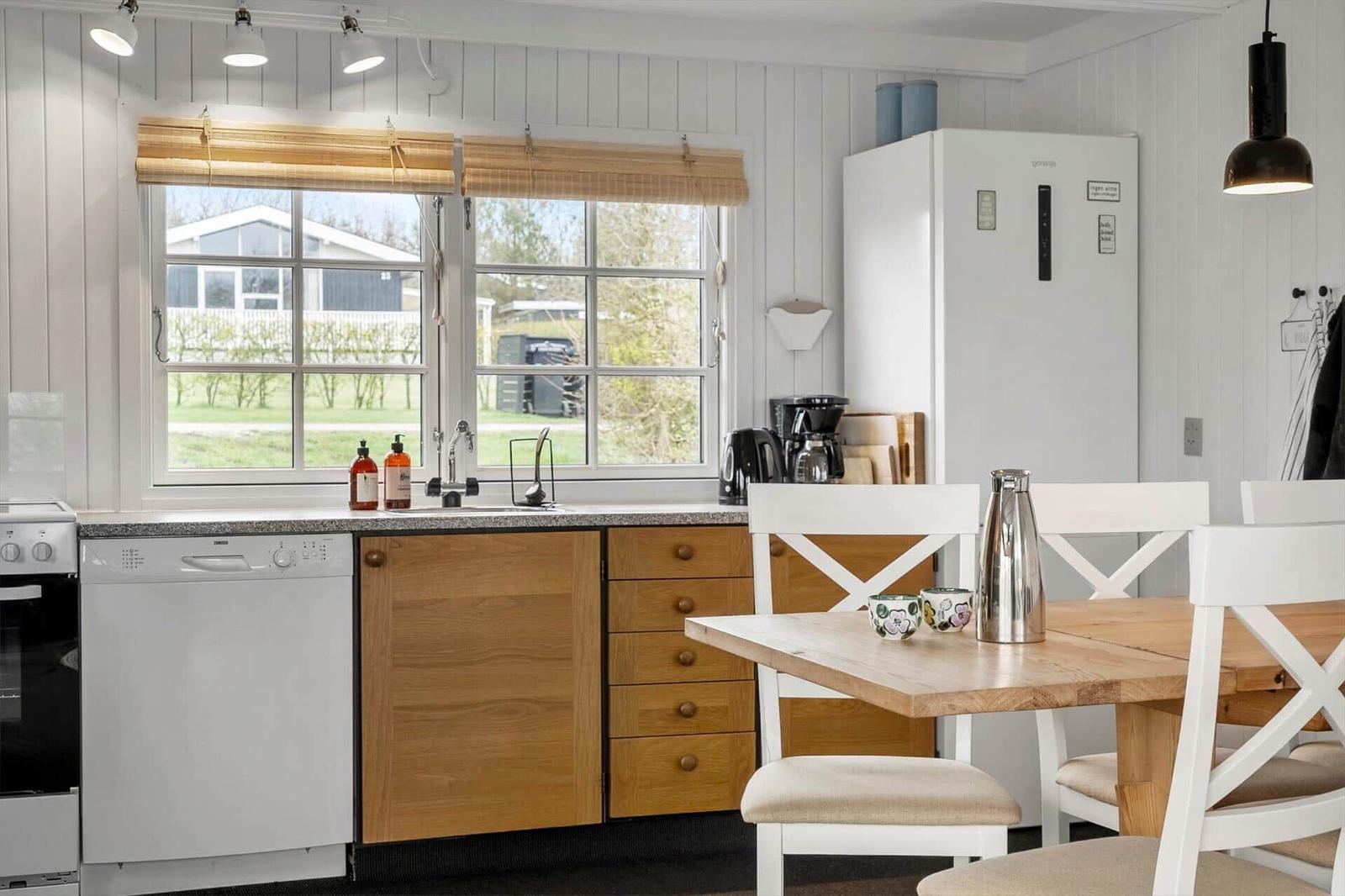 Küche mit Holzschränken, Waschmaschine und Tisch mit Stühlen. Fenster mit Blick auf Grünfläche.
