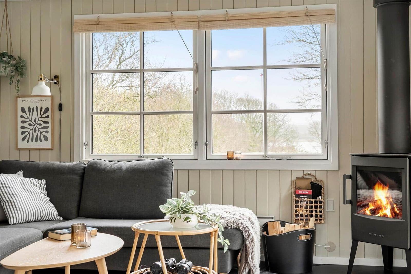 Ein Wohnzimmer mit grauem Sofa, Kamin und großem Fenster mit Blick auf Bäume.